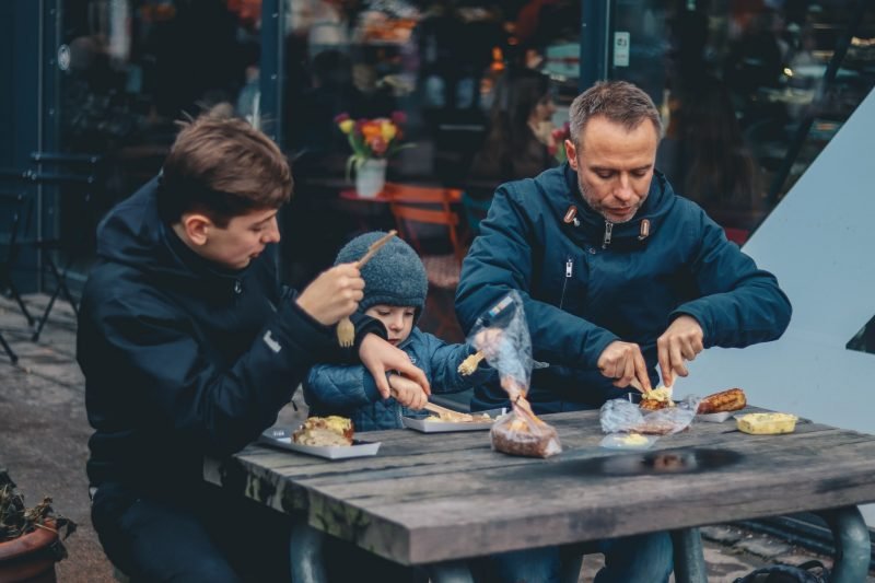 Dos hombres y un niño sentados en una banca satisfaciendo la necesidad básica de comer.